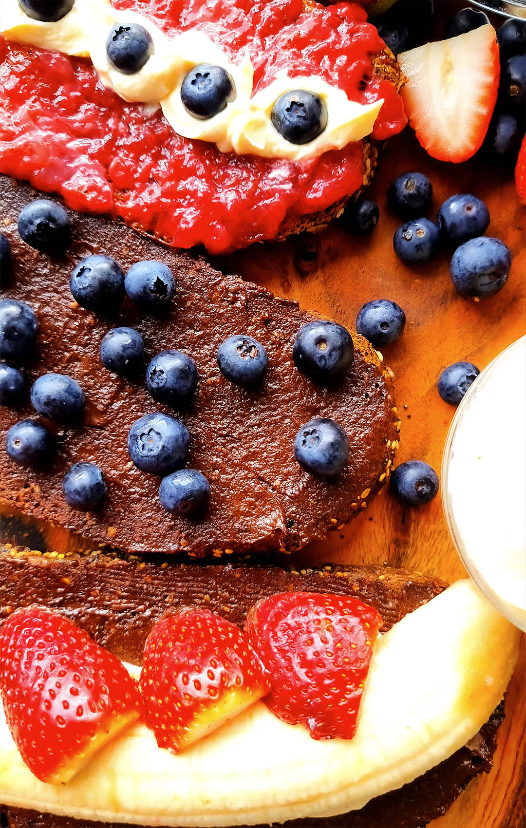 Close-up of thick, creamy Healthy Homemade Choco-Nuts Butter spread on dark bread — richly layered and topped with fresh blueberries, strawberries, and banana slices. The texture of the nut butter is smooth and glossy, highlighting its rich, chocolatey depth.