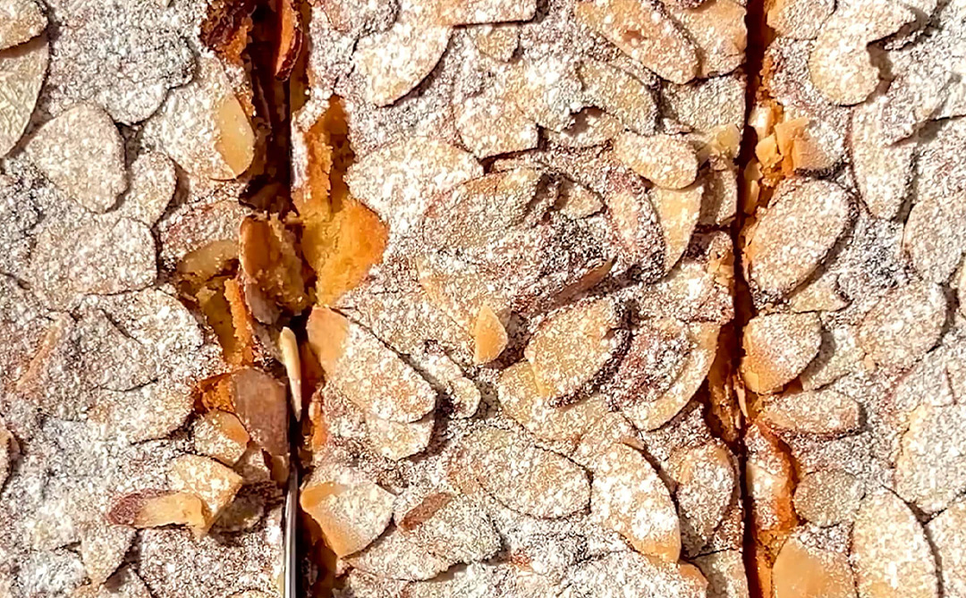 Close-up of sweetener-dusted bars being sliced, showing the frangipane filling and crispy almond topping breaking apart with a golden crunch. Low-Carb and Paleo dessert.