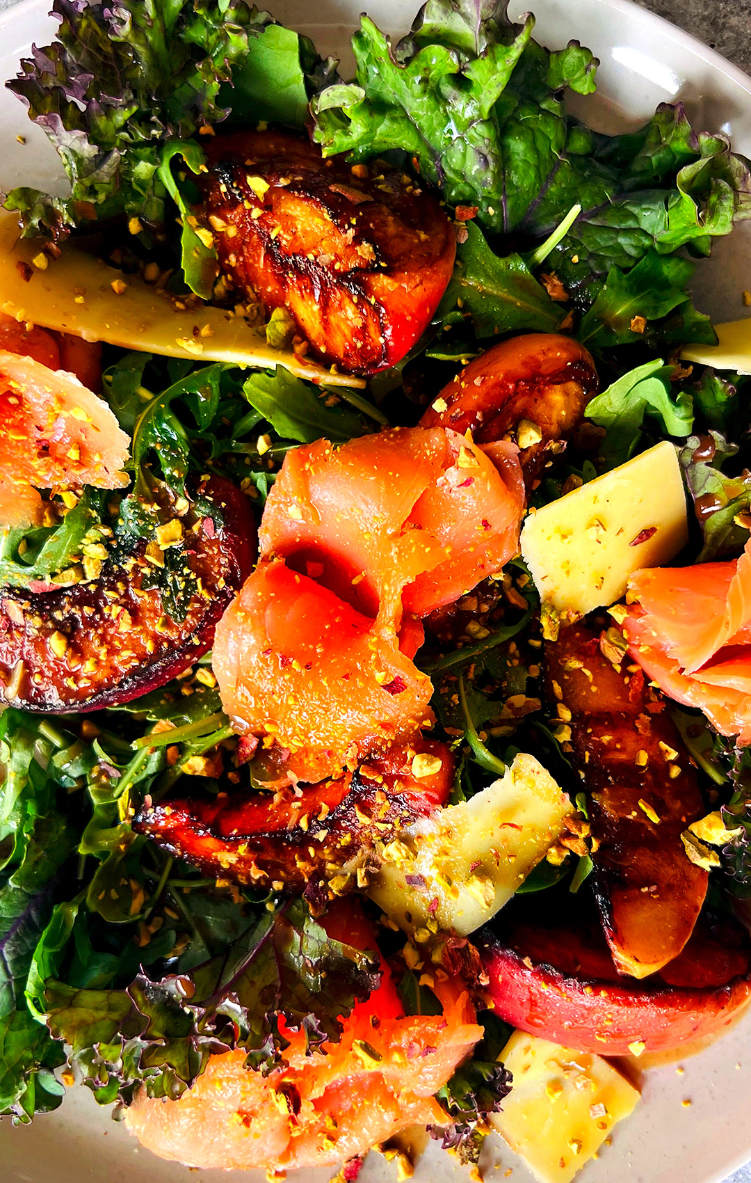 "Zoomed-in view of caramelized peach, smoked salmon, and curly green kale salad topped with chopped pistachios, shaved smoked Cheddar, and honey Dijon balsamic vinaigrette on a serving plate, showing how satisfying and delicious it is! A protein-rich salad perfect for lunch