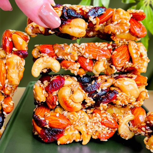 Stack of pan-made nut and seed bars with one bar being lifted by hand to show crisp, crunchy texture. Healthy no-bake snack made with almonds, cashews, sesame seeds, and natural sweetener. Paleo, gluten-free, and refined sugar-free.