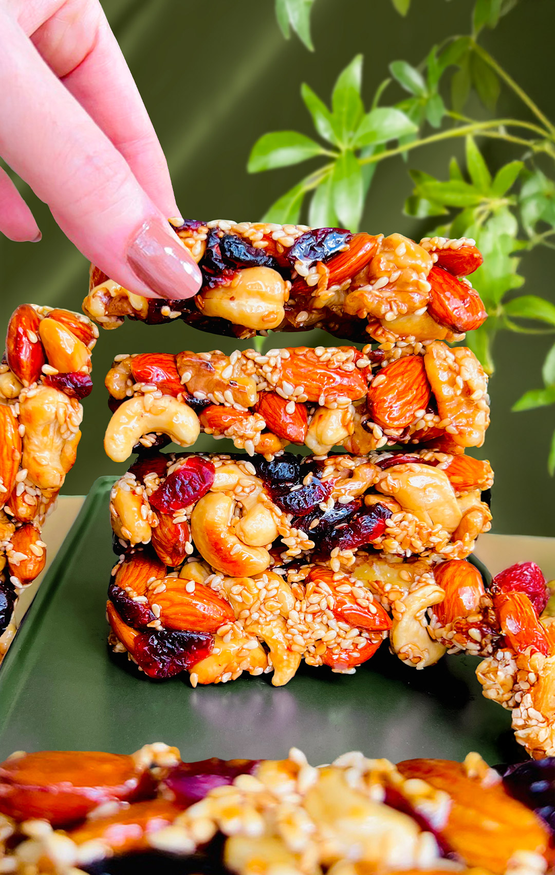 Stack of pan-made nut and seed bars with one bar being lifted by hand to show crisp, crunchy texture. Healthy no-bake snack made with almonds, cashews, sesame seeds, and natural sweetener. Paleo, gluten-free, and refined sugar-free.