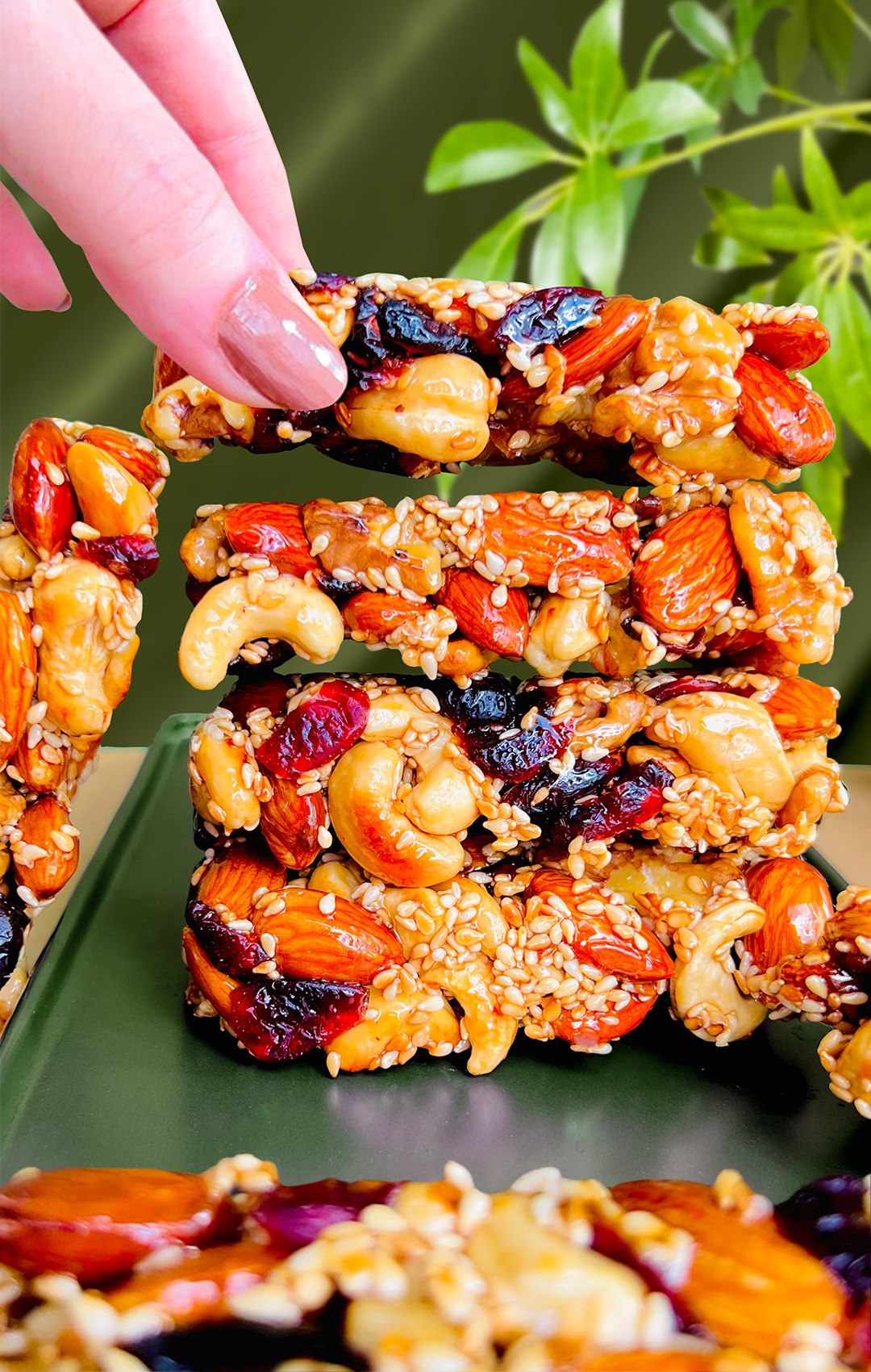 Stack of pan-made nut and seed bars with one bar being lifted by hand to show crisp, crunchy texture. Healthy no-bake snack made with almonds, cashews, sesame seeds, and natural sweetener. Paleo, gluten-free, and refined sugar-free.