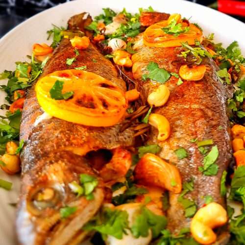 Close-up of the golden, crispy-skinned ocean trout showing flaky texture, smooth roasted garlic, toasted cashews, and red chilli, topped with roasted lemon slices and fresh parsley.