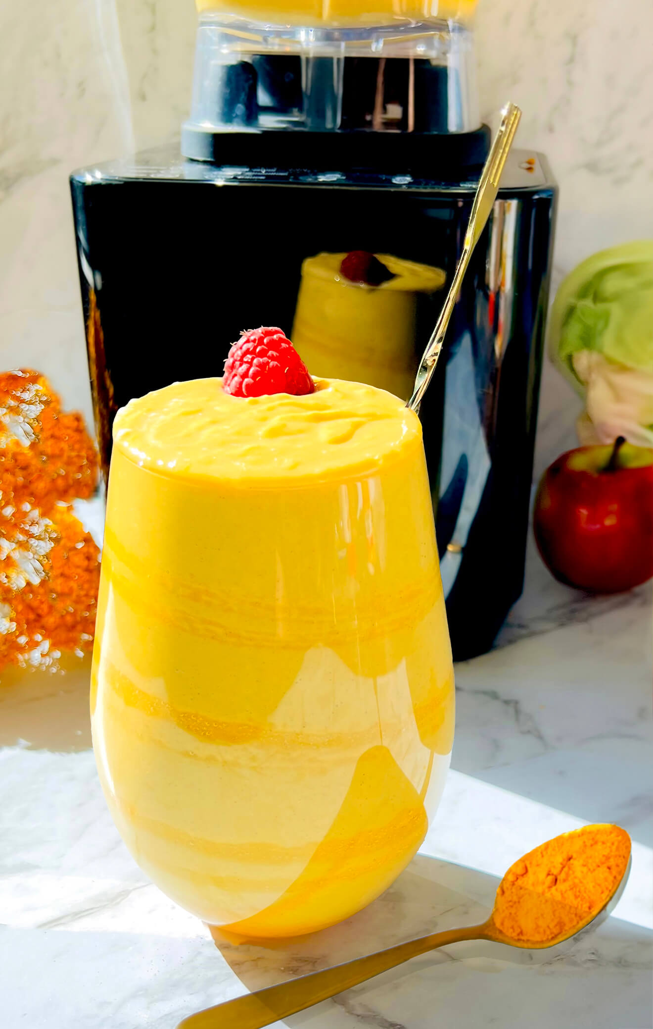 Creamy golden glow smoothie in a glass under sunlight, with a spoonful of turmeric, cauliflower, and apple in the background. Perfect for anti-inflammatory diet, perfect for breakfast. Made with cauliflower, Greek yogurt, turmeric powder, ginger, almond milk, and tahini, honey, pumpkin spice, cinnamon.