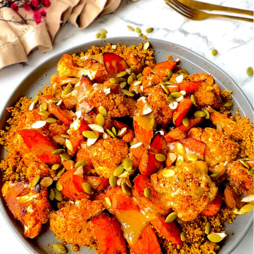 Smoky roasted cauliflower and carrots, caramelised to perfection, served over golden fluffy quinoa infused with turmeric and cinnamon, drizzled with creamy rich tahini sauce, and presented on a plate next to cutlery and flowers.
