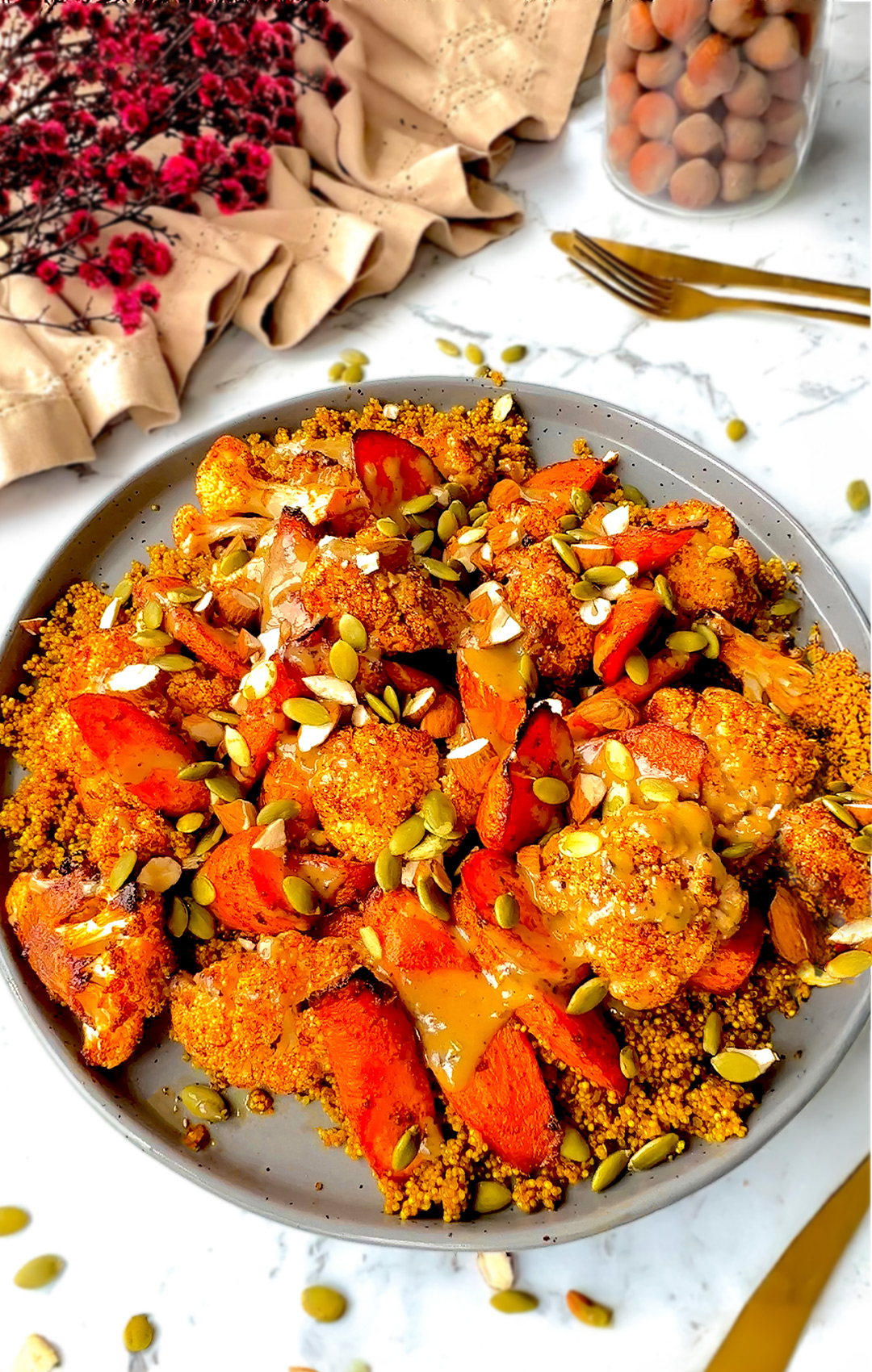 Smoky roasted cauliflower and carrots, caramelised to perfection, served over golden fluffy quinoa infused with turmeric and cinnamon, drizzled with creamy rich tahini sauce, and presented on a plate next to cutlery and flowers.