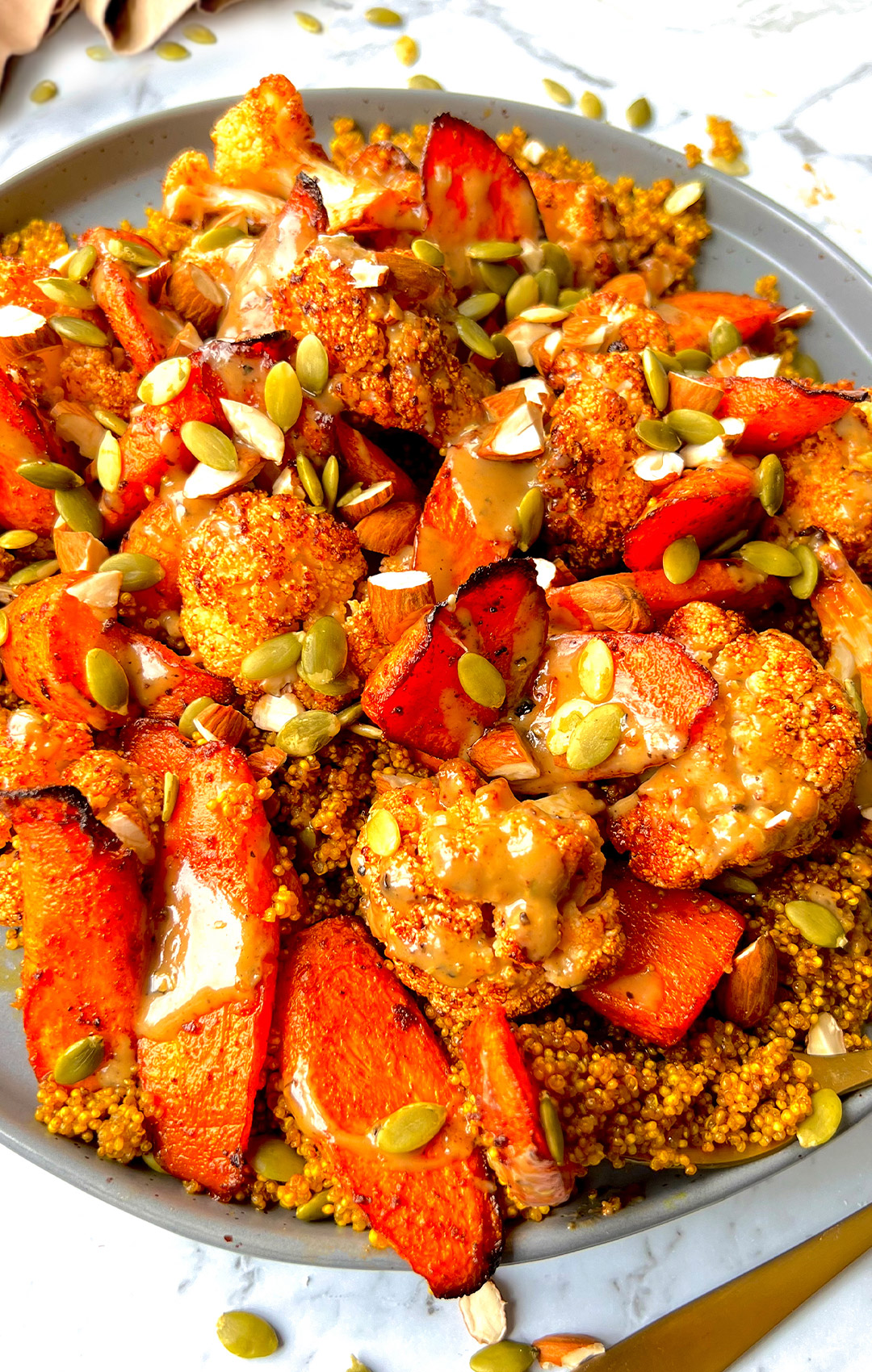 Close-up of fluffy golden quinoa, smoky roasted cauliflower and carrots caramelised to perfection, with crunchy almonds, pumpkin seeds, and a rich tahini drizzle next to cutlery and flowers.