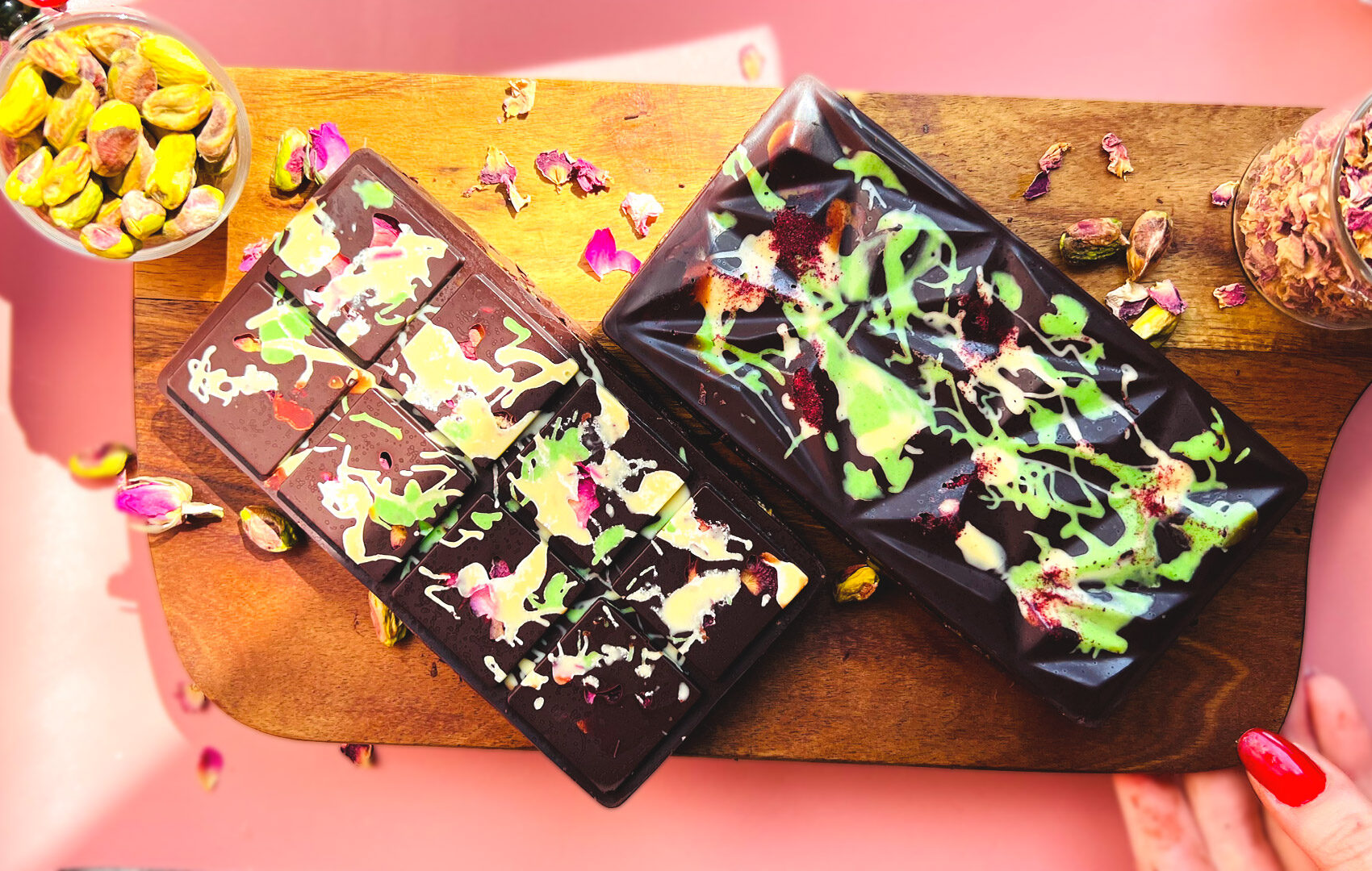 Two Healthy Dubai Chocolate Bars on a wooden board, one decorated with raspberry powder and white-green drizzle, the other with rose petals and pistachio accents. Inside filling made from pistachio-tahini cream and toasted coconut for a rich multi-textural bite. Healthy, gluten free, low carb, paleo, refined sugar free, keto option.