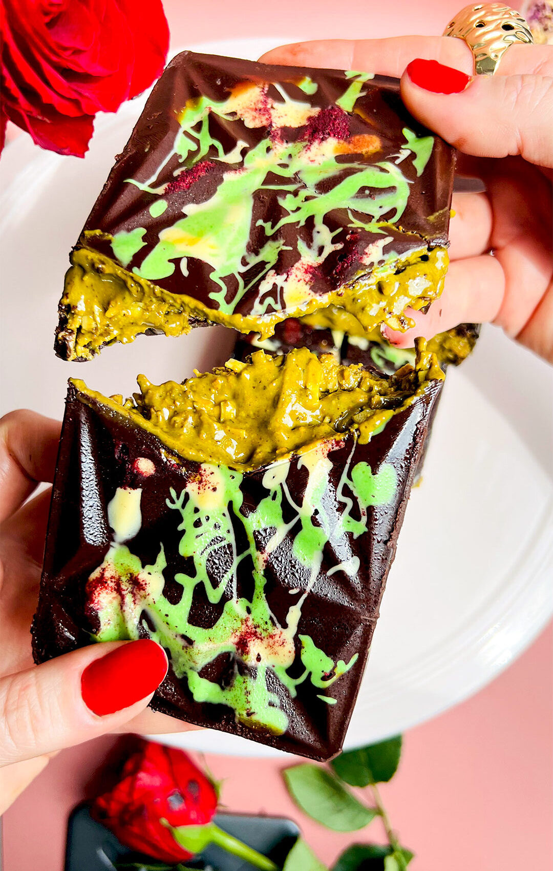 Hands breaking a Healthy Dubai Chocolate Bar in half, revealing vibrant green, gooey pistachio-tahini and toasted coconut filling under a glossy shell decorated with white and green drizzle. Creamy, crunchy, and gooey texture with rose petals for a boutique finish. Low carb, gluten free, paleo, refined sugar free, keto option.