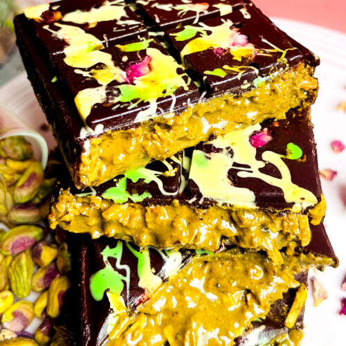 Stacked halves of Healthy Dubai Chocolate Bars with creamy pistachio-tahini and toasted coconut filling oozing out, topped with glossy dark chocolate decorated with rose petals and colourful drizzle. Unique multi-textural experience that is creamy, smooth, crunchy, and gooey. Healthy, refined sugar free, gluten free, low carb, keto and paleo friendly.