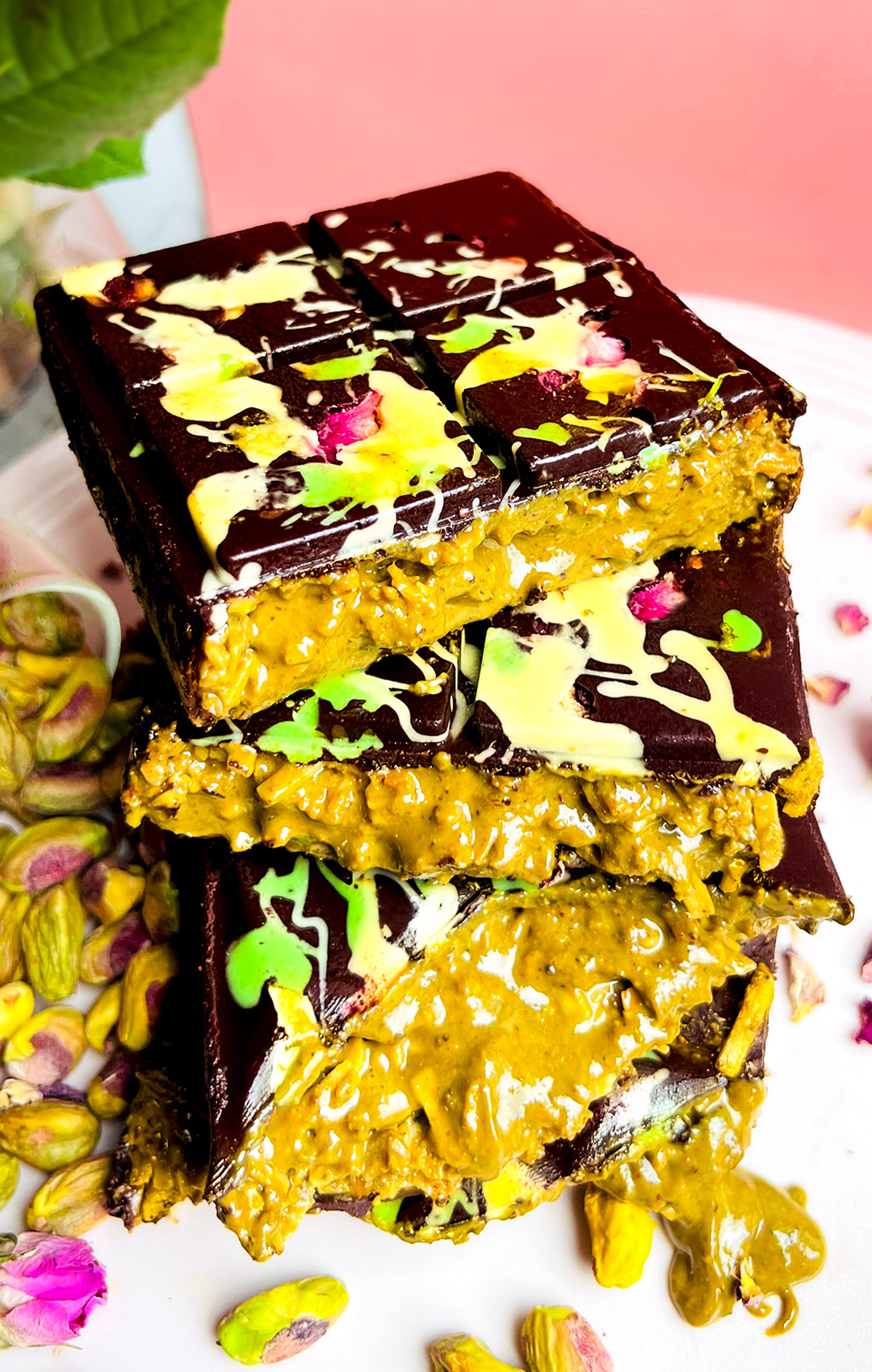 Stacked halves of Healthy Dubai Chocolate Bars with creamy pistachio-tahini and toasted coconut filling oozing out, topped with glossy dark chocolate decorated with rose petals and colourful drizzle. Unique multi-textural experience that is creamy, smooth, crunchy, and gooey. low carb chocolate bars with pistachio filling are Healthy, refined sugar free, gluten free, low carb, keto and paleo friendly.