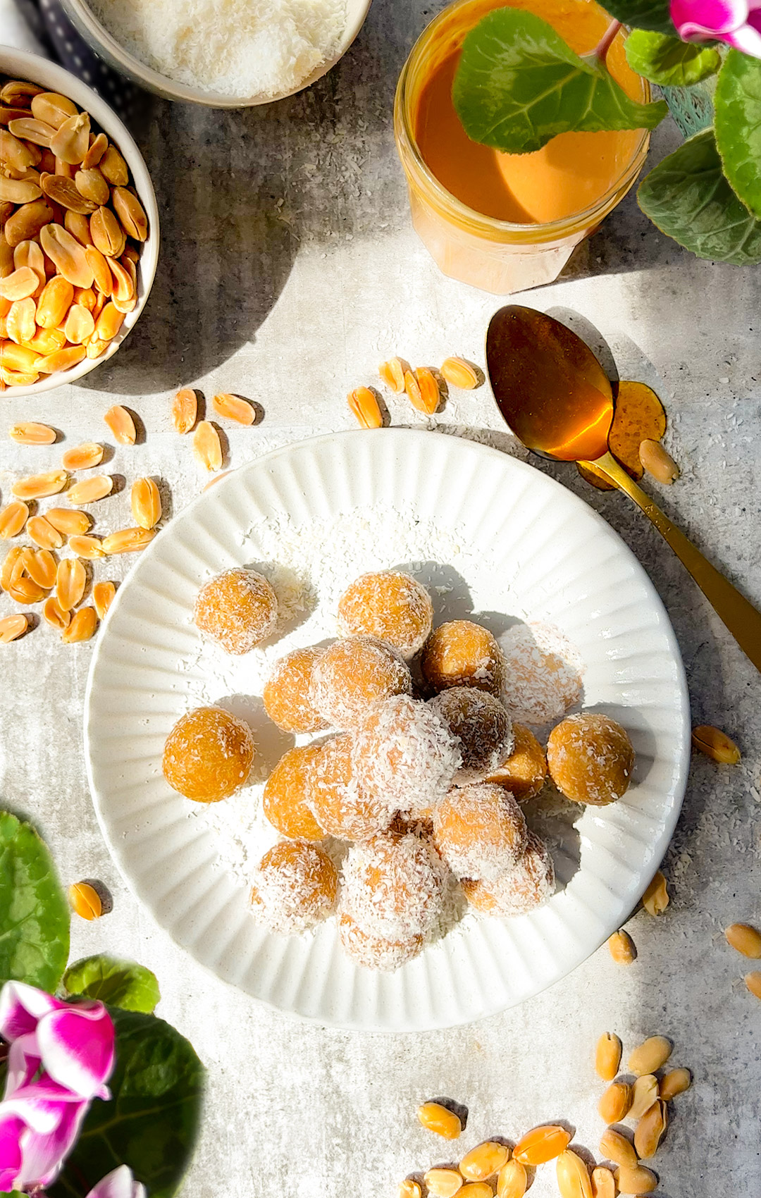 White plate of homemade peanut butter coconut energy balls coated in shredded coconut, surrounded by peanuts and a glass of peanut butter drink. Easy 5-ingredient keto, paleo, gluten free snack.
