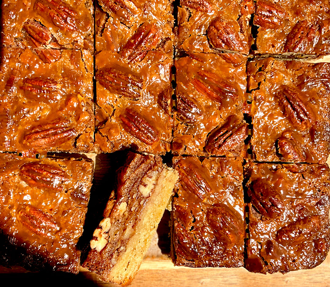Overhead view of healthy Healthy Caramel Pecan Pie Bars cut into squares with one slice turned on its side to show the layers, gooey chewy caramel pecan topping with crackly shiny surface, crunchy toasted pecans and a thick buttery almond flour shortbread base,