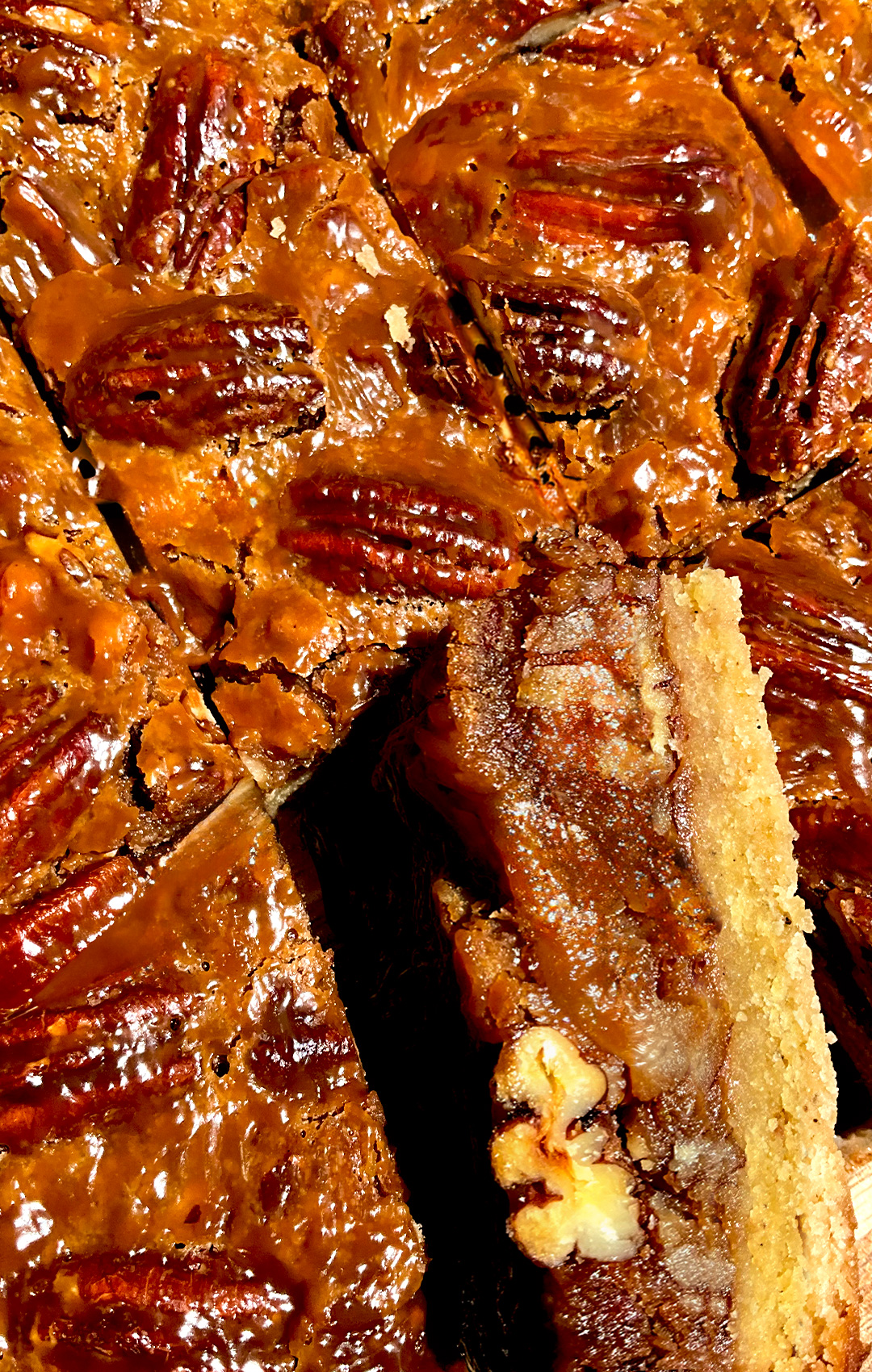 © Healthy Food by Annie – All rights reserved. No reuse without permission. Tray of healthy caramel pecan pie bars with one slice turned on its side to show gooey soft caramel pecan layer and soft almond flour shortbread crust, shiny crackled top with toasted pecans, paleo, gluten free, grain free, refined sugar free lower carb dessert. caramel pie layer made with maple syrup and brown monk fruit, packed with toasted pecans on top of a buttery almond flour shortbread base. For keto option Swap maple syrup for sugar free maple syrup