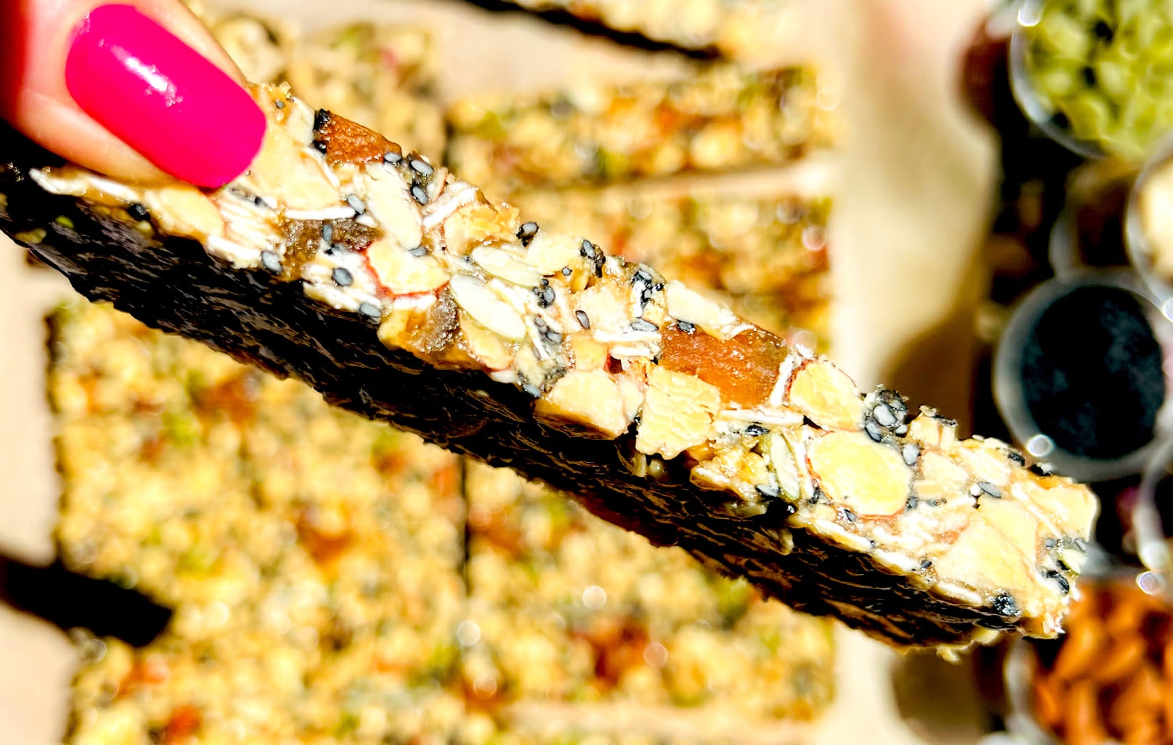 close up finger holding a sliced oat, honey and tahini granola bar showing texture, and wholesome superfood ingredients like seeds, nuts and dried apricot, no refined sugar, healthy gluten free snack for grab and go breakfast or meal prep perfect for pre and post workout