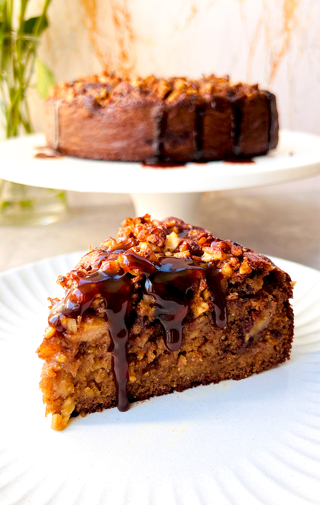 paleo sticky apple pudding cake serves warm, featuring a soft sticky pudding texture, caramelised apples and walnuts topping, with caramelise crispy edges and rich coconut caramel sauce drizzle. Its made of wholesome ingredients, almond flour, coconut sugar, Healthy, Gluten-free, dairy-free and refined sugar-free. At background whole cake with crispy caramelise edges Thick, glossy caramel sauce dripping down the sides looks divine!