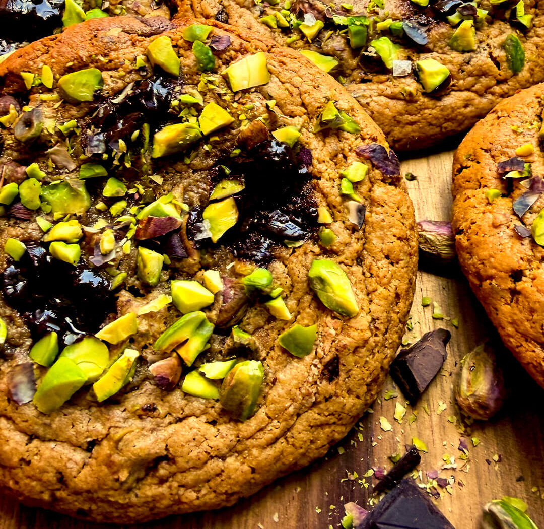 Closeup Detailed texture of pistachio chocolate chunk cookie showing caramelised crispy edges, soft centre and glossy melted dark chocolate pieces.
