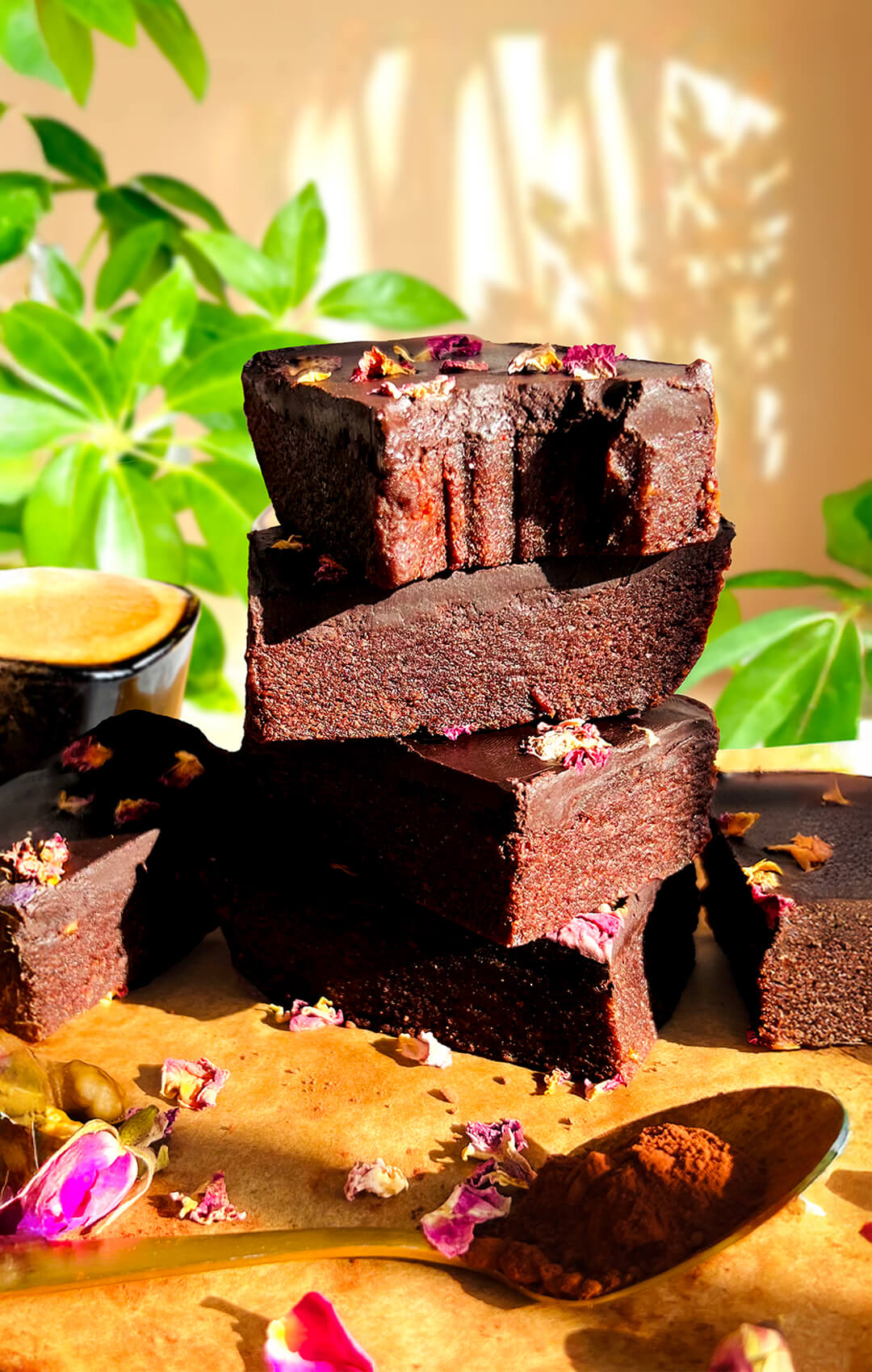 Stacked healthy no-bake date brownies with a bite taken from the top piece, showing a gooey fudgy chocolate center, glossy dark chocolate ganache, rose petals, walnuts, and cocoa powder, a naturally sweetened paleo and gluten-free dessert with no refined sugar, quick and easy recipe and perfect for meal prep.