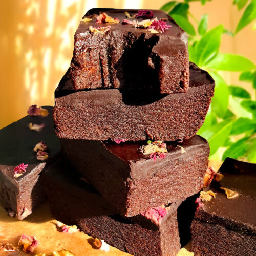 Close-up of stacked healthy no-bake date brownies with smooth glossy chocolate ganache and rose petals, showing their dense fudgy layers and rich chocolatey texture, naturally sweetened with dates and honey, paleo, gluten-free, and made with walnuts, cocoa, tahini, and dark chocolate.