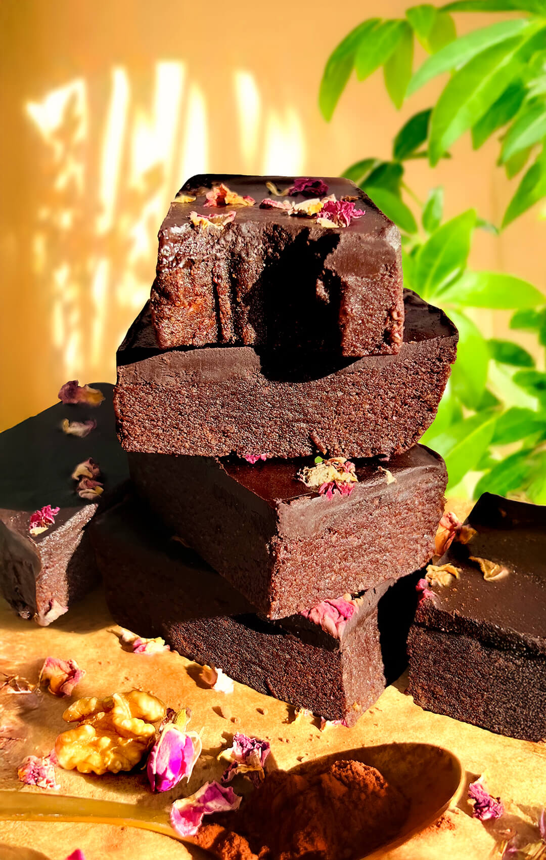 Close-up of stacked healthy no-bake date brownies with smooth glossy chocolate ganache and rose petals, showing their dense fudgy layers and rich chocolatey texture, naturally sweetened with dates and honey, paleo, gluten-free, and made with walnuts, cocoa, tahini, and dark chocolate.