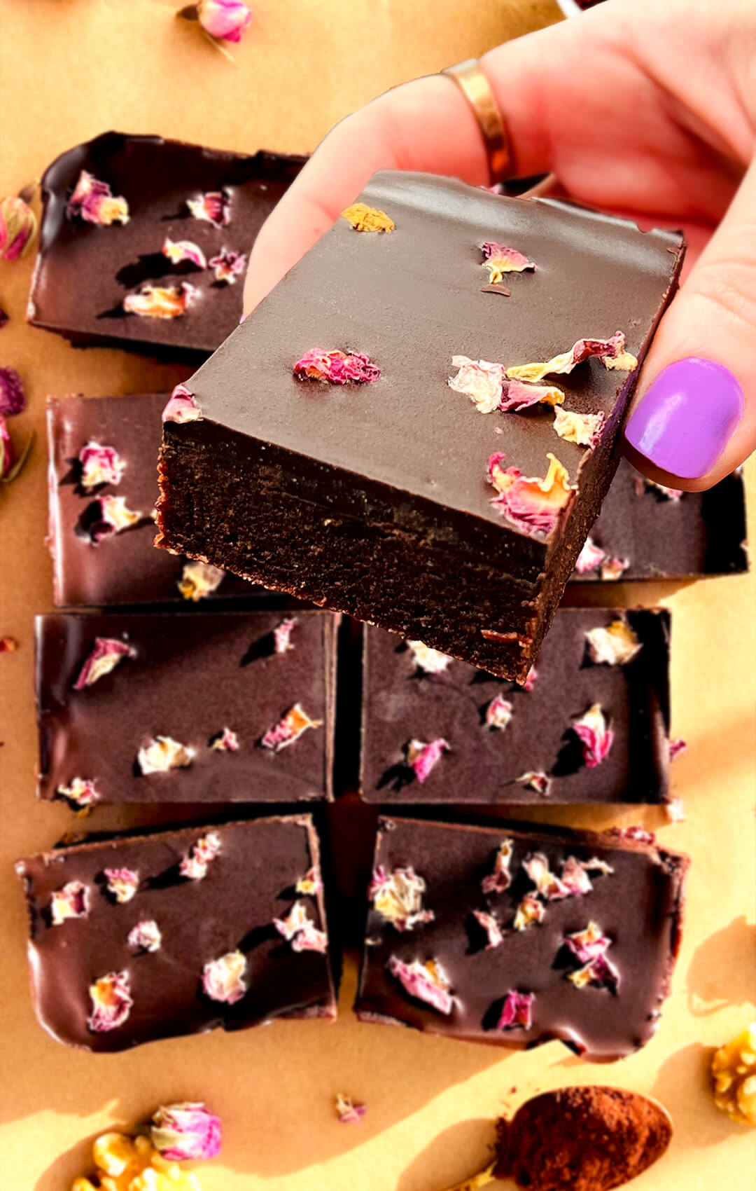 Hand holding a square of healthy no-bake date brownie with a smooth glossy chocolate topping and rose petals, showing its thick fudgy texture and clean layers, naturally sweetened, paleo, gluten-free, and made with walnuts, cocoa, tahini, and dark chocolate. These are so easy to make in 15 minutes and perfect for meal prep and for afternoon coffee.