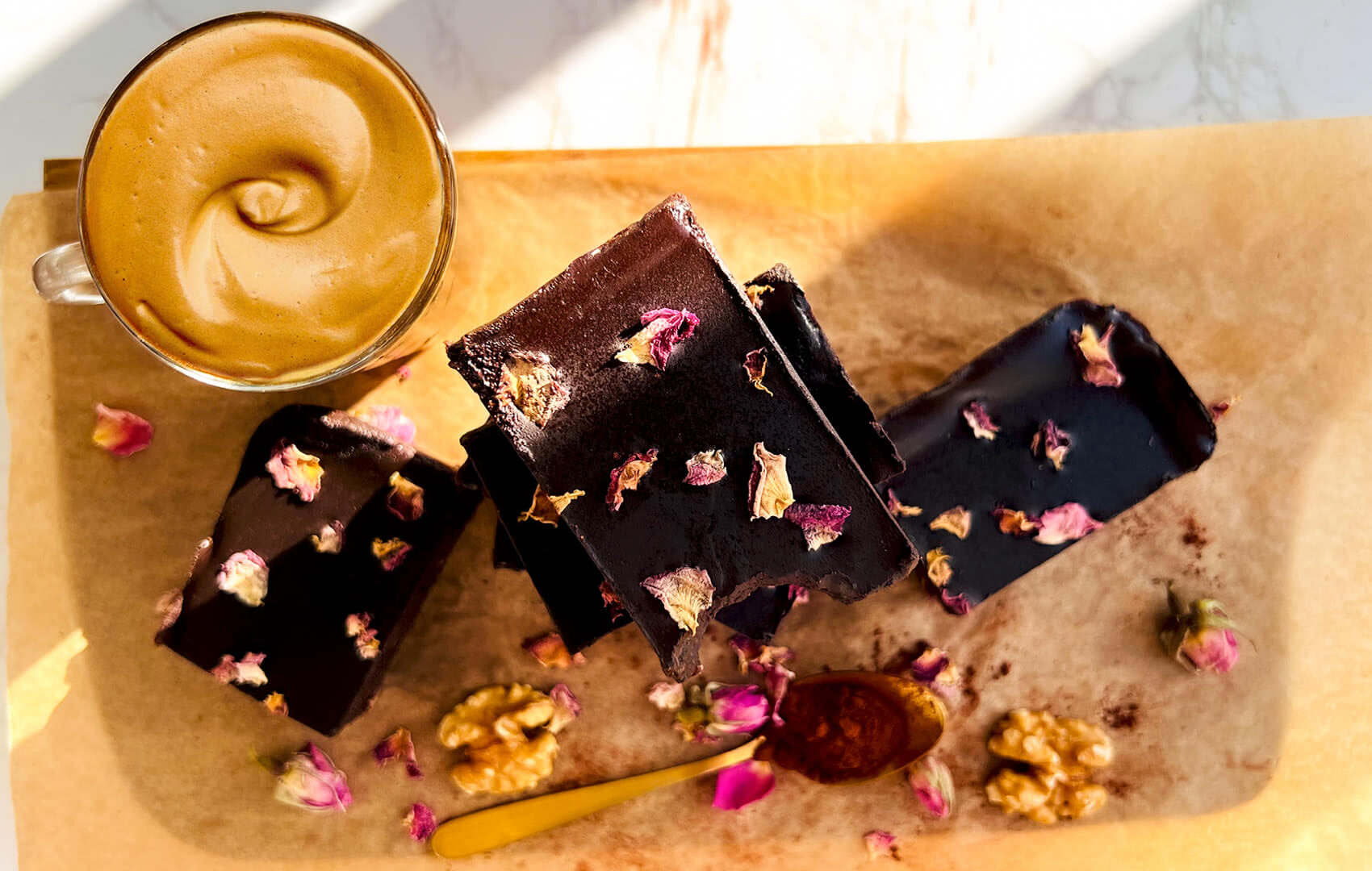 Overhead view of healthy no-bake date brownies served with coffee on parchment paper, topped with glossy dark chocolate ganache and rose petals, rich and fudgy, naturally sweetened, paleo, gluten-free, and perfect for a sweet snack or elegant dessert. Quick and easy to make, for guests, afternoon tea, also perfect for meal prep, and pre workout.