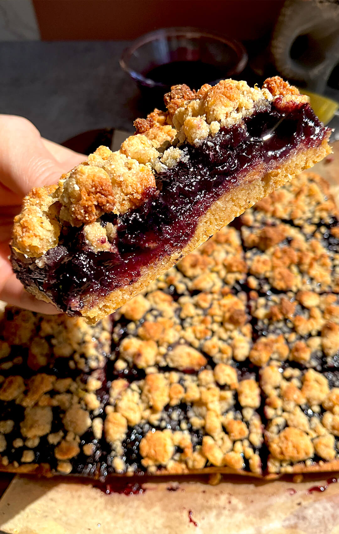 A slice of blueberry crumble bar being lifted to show the bakery-style layers, soft almond and coconut flour base, thick jammy blueberry sauce with whole berries, and a buttery golden crumble top, paleo, gluten-free and dairy-free, refined sugar-free with vegan and lower sugar options.