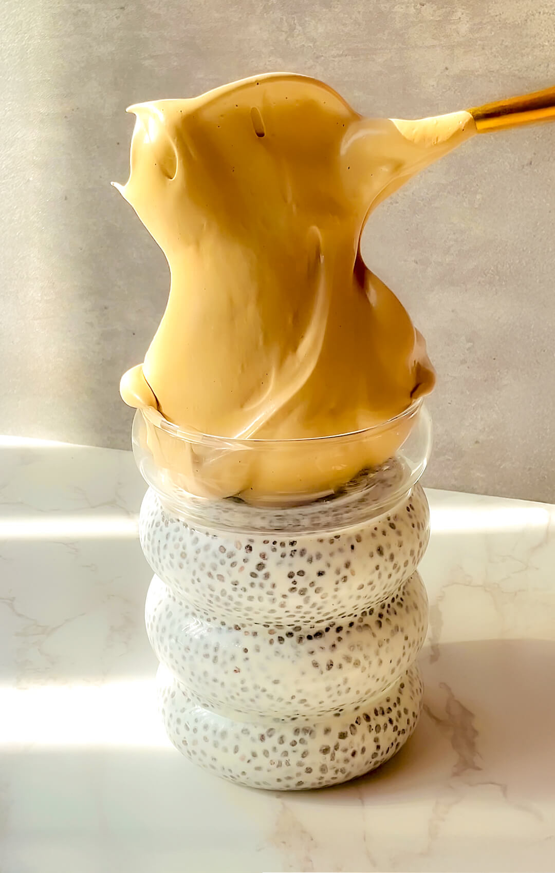 Thick, glossy, stretchy whipped honey dalgona coffee being lifted from a spoon over creamy chia pudding, showing a velvety stable texture that holds its shape beautifully, a healthy mix of morning coffee and breakfast that is quick and easy to make in 5 minutes, naturally sweetened, paleo, gluten-free, dairy-free, refined sugar-free, and perfect for breakfast or an afternoon treat.