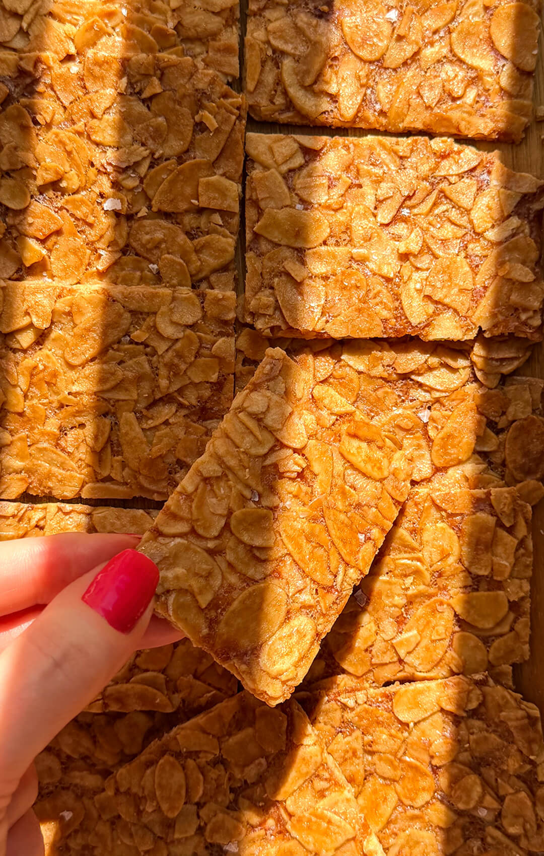 Hand holding a thin almond crisp cookie over a tray of baked cookies, showing the very thin crisp texture, deeply golden sliced almonds, and delicate crackly finish. a healthier version of Almond Florentines. These sugar-free almond crisp cookies are flourless, keto, gluten-free, vegetarian, paleo option and made with simple ingredients for a quick and easy almond cookie recipe with a light snap in every bite. healthier version of Almond Florentines