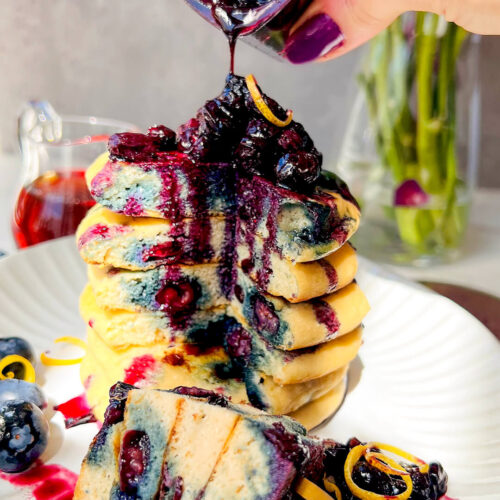 Healthy blueberry sauce being poured over a stack of fluffy paleo blueberry pancakes on a white plate, with a sliced pancake in front and a fork next to the stack showing the soft, thick, pillowy texture, juicy blueberries, naturally light sweetness, and a healthy gluten-free dairy-free grain-free breakfast perfect for weekend brunch.