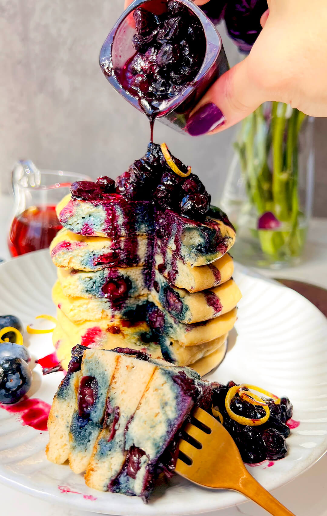Healthy blueberry sauce being poured over a stack of fluffy paleo blueberry pancakes on a white plate, with a sliced pancake in front and a fork next to the stack showing the soft, thick, pillowy texture, juicy blueberries, naturally light sweetness, and a healthy gluten-free dairy-free grain-free breakfast perfect for weekend brunch.