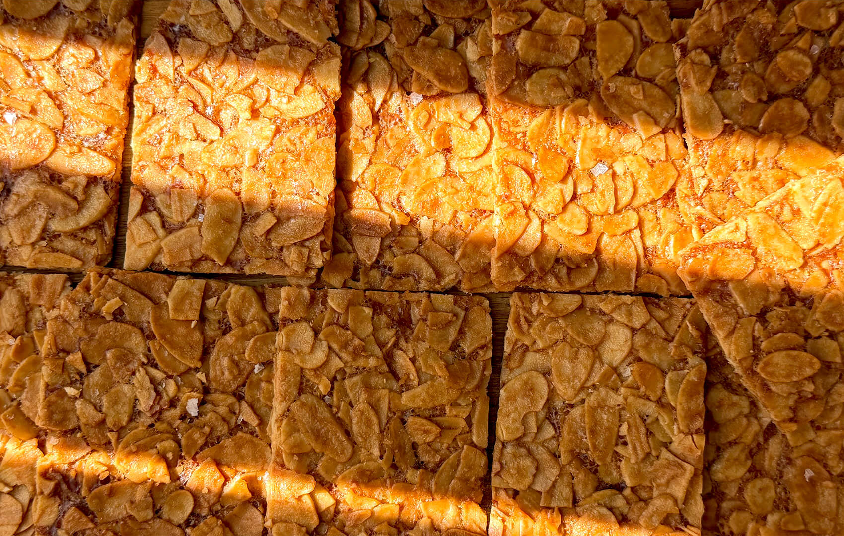 Close-up of baked almond crisp cookies cut into rectangles lineup, showing a very thin golden crisp texture, deeply golden sliced almonds, and delicate crackly finish under sun light. These sugar-free flourless almond cookies are crunchy, flavourful, and incredibly easy to make at home, with keto, gluten-free, vegetarian, and paleo option appeal. made with sliced almonds, egg white, golden monkfruit sweetener, unsalted butter, vanilla, and almond extract