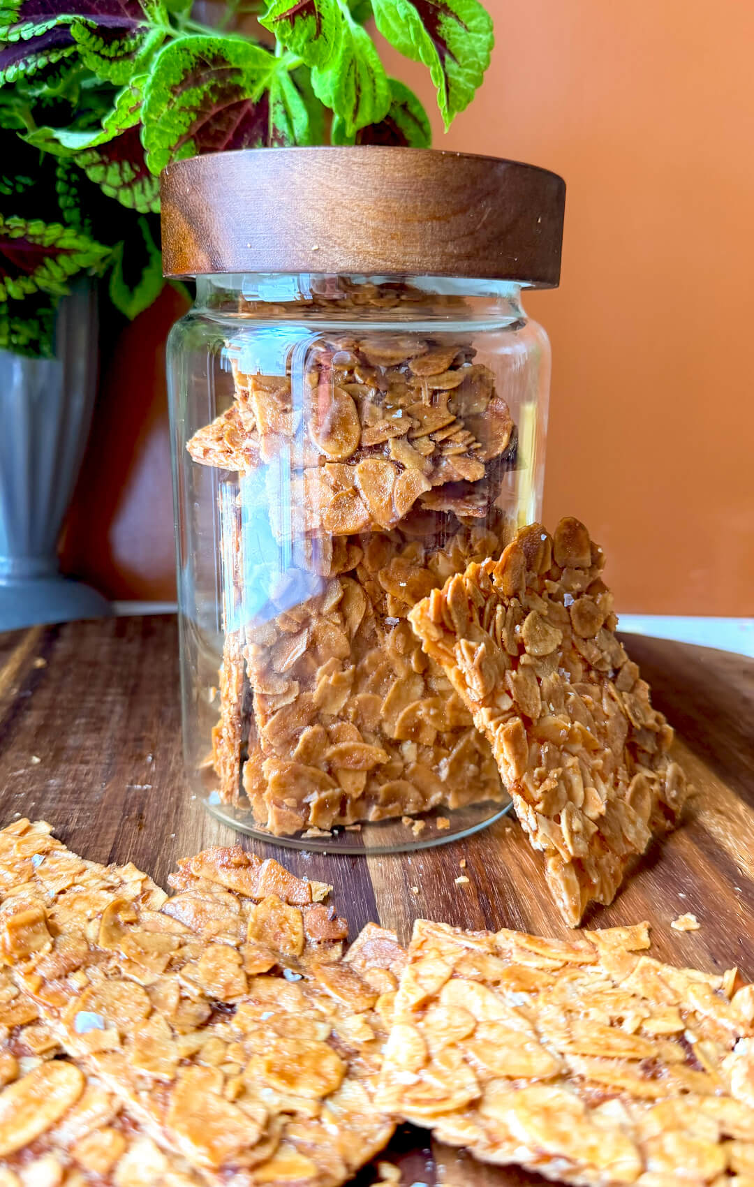 Sugar-free almond crisp cookies stored in a glass jar on a wooden board, with extra cookies arranged around the jar for a make-ahead and meal prep friendly serving idea, and showing how thin and crispy they are. These flourless almond cookies are very thin, crisp, deeply golden, and made with simple ingredients can made easily at home, with keto, gluten-free, vegetarian, and paleo option appeal. made with sliced almonds, egg white, golden monkfruit sweetener, unsalted butter, vanilla, and almond extract for paleo replace butter with ghee.