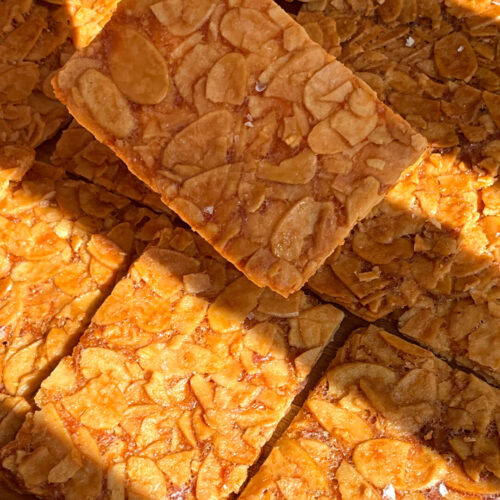 Hand holding a thin golden almond crisp cookie, with a lineup of baked almond crisp cookies in the background with very thin crisp texture. These sugar-free flourless almond cookies are crunchy, quick and easy to make at home, with a delicate crackly finish, keto, gluten-free, vegetarian, and paleo option replacing butter with ghee. healthier version of Almond Florentines. made with sliced almonds, egg white, golden monkfruit sweetener, unsalted butter, vanilla, and almond extract.