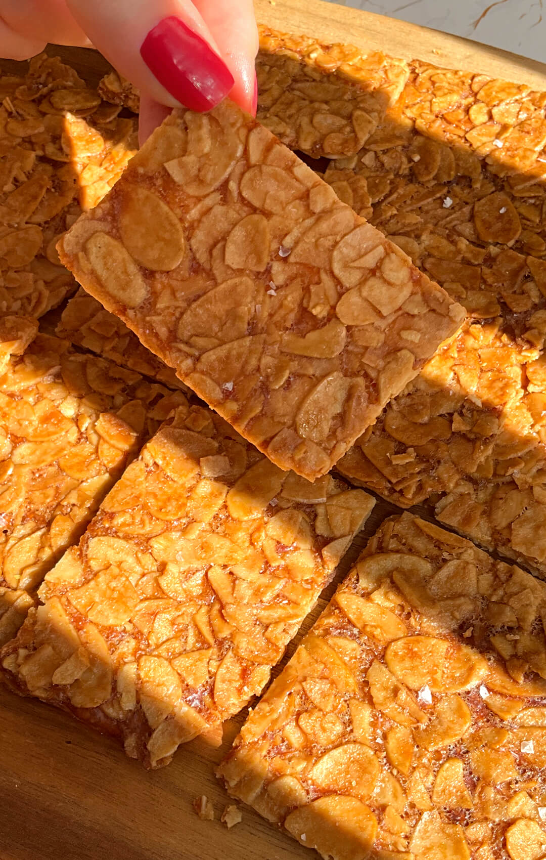 Hand holding a thin golden almond crisp cookie, with a lineup of baked almond crisp cookies in the background with very thin crisp texture. These sugar-free flourless almond cookies are crunchy, quick and easy to make at home, with a delicate crackly finish, keto, gluten-free, vegetarian, and paleo option replacing butter with ghee. healthier version of Almond Florentines. made with sliced almonds, egg white, golden monkfruit sweetener, unsalted butter, vanilla, and almond extract.