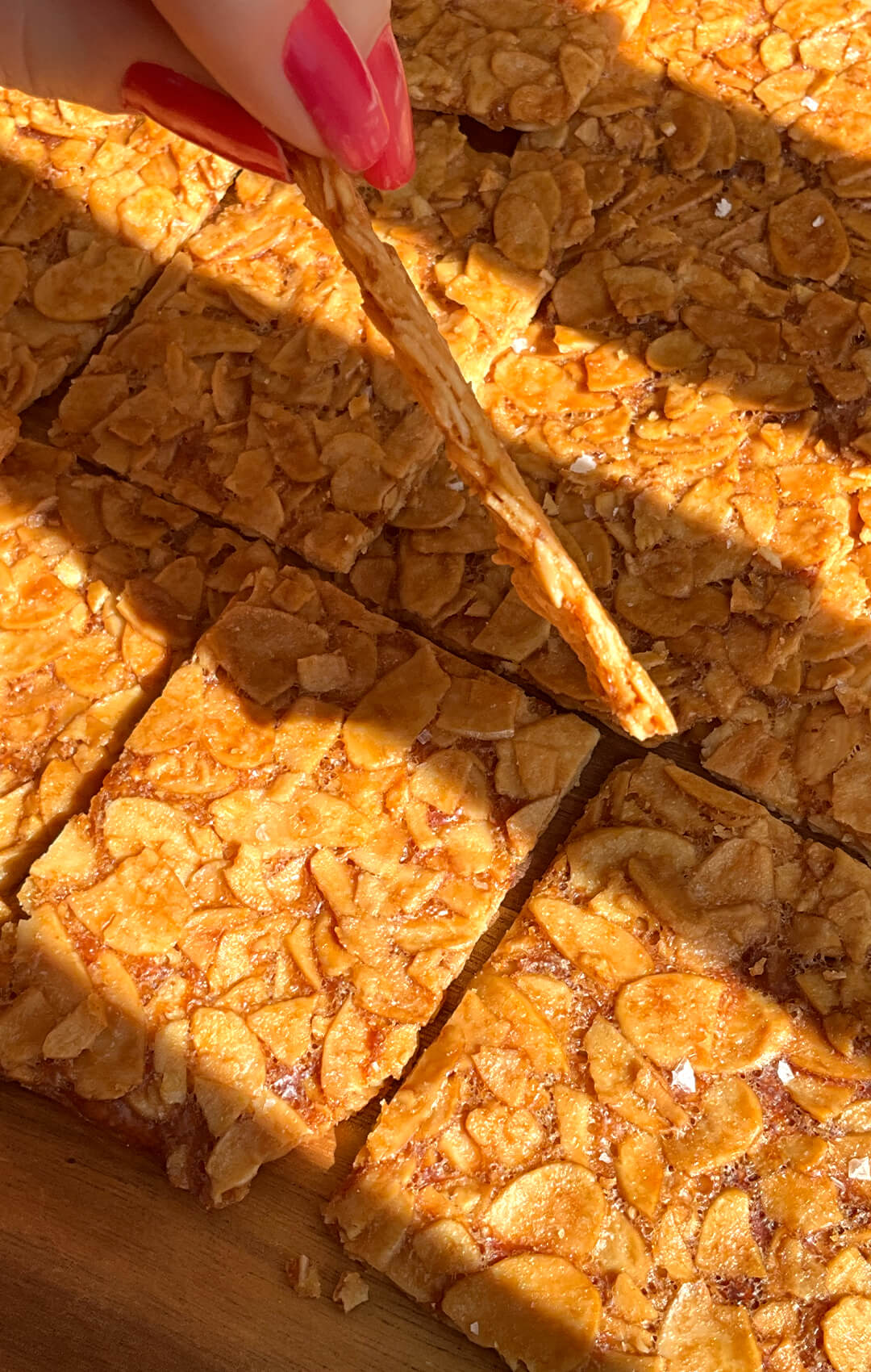Close-up of a thin golden almond crisp cookie held by hand, with golden almond crisp cookies in the background. showing the deeply golden sliced almonds and delicate crackly finish. These sugar-free flourless almond cookies are made with simple ingredients for a thin crispy almond cookie recipe with keto, gluten-free, vegetarian, and paleo option appeal. healthier version of Almond Florentines