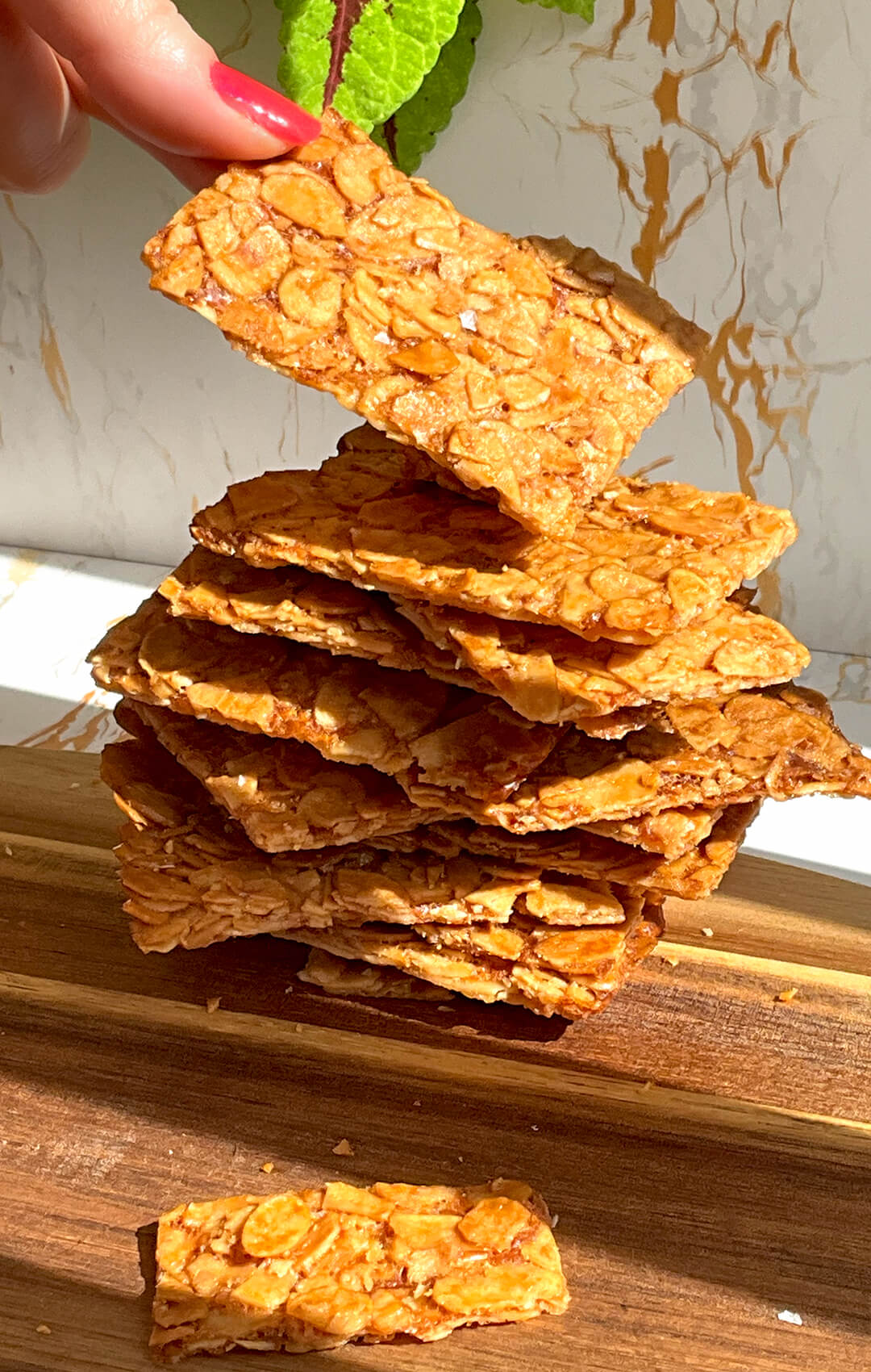Hand placing a thin golden almond crisp cookie on top of a stack of baked almond crisp cookies on a wooden board.showing the deeply golden sliced almonds, very thin crisp texture, and delicate crackly finish. These sugar-free flourless almond cookies are quick and easy to make with simple ingredients, with keto, gluten-free, vegetarian, and paleo option appeal.