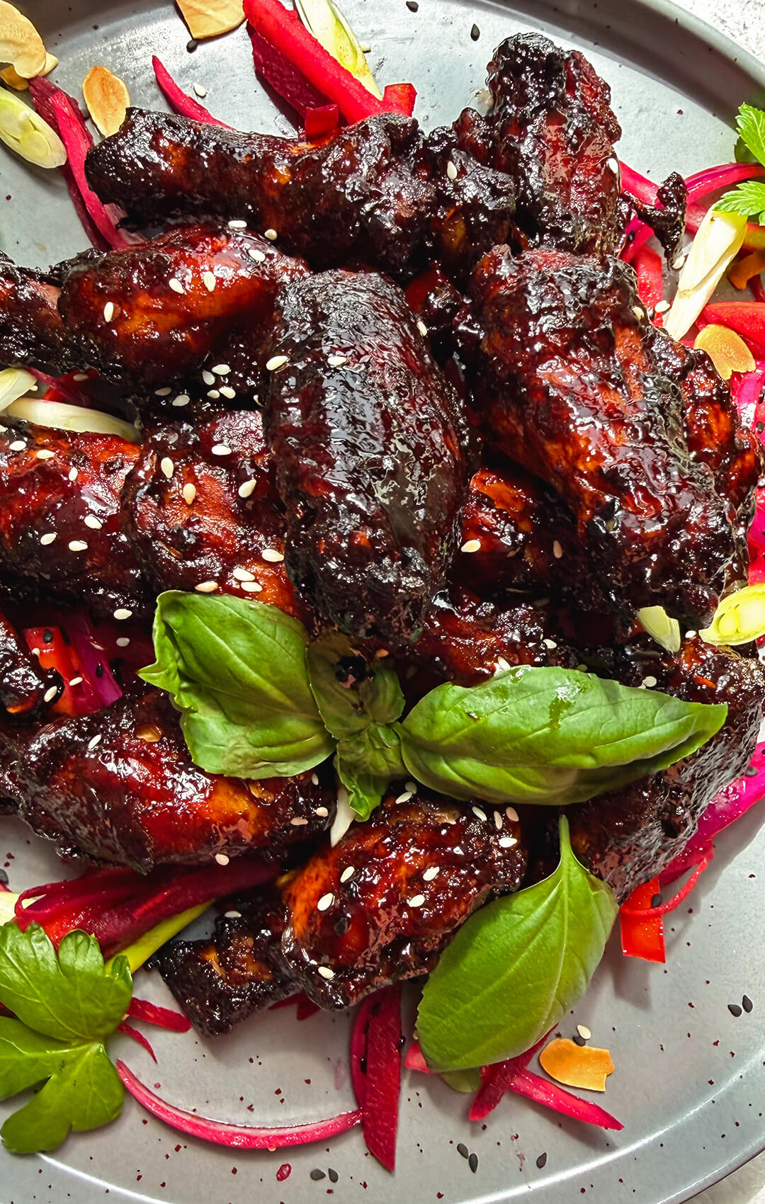 Crispy sticky honey soy air fryer chicken wings served on a plate with a dark red secret glaze, sesame seeds, fresh herbs, pink salad, and toasted almonds. These healthy air fryer chicken wings are naturally sweetened with honey, dairy-free, high-protein, smoky, sweet, savoury, sticky, glossy, caramelised, and full of tender juicy flavour.