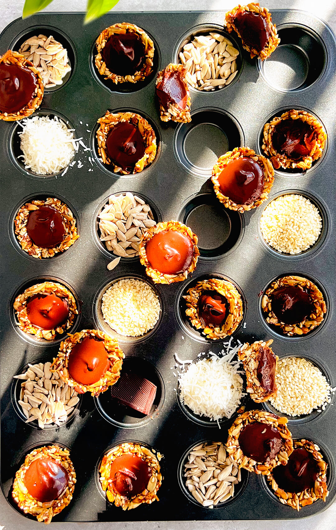 No-Oven Chocolate Tahini Seed Cups arranged in a muffin tray, made with golden crunchy toasted sunflower seeds, sesame seeds, shredded coconut, and honey, then filled with silky, glossy dark chocolate tahini. These easy pan-made seed cups are ready in under 30 minutes, paleo, gluten-free, nut-free, dairy-free, oil-free, refined sugar-free, delicious, wholesome, and perfect for a healthy chocolate snack, lunch box treat, or easy meal prep.