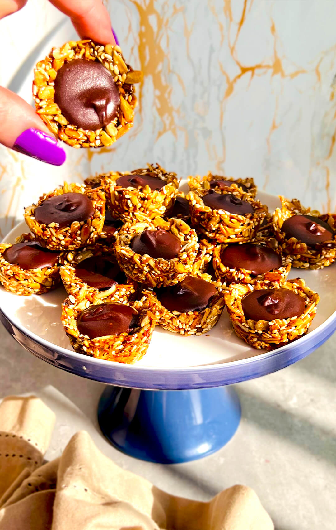 Healthy No-Oven Chocolate Tahini Seed Cups piled on a cake stand, with crunchy golden seed cups made from sunflower seeds, sesame seeds, shredded coconut, and honey, filled with silky, glossy dark chocolate tahini. These wholesome chocolate cups are gluten-free, nut-free, dairy-free, oil-free, refined sugar-free, paleo-friendly, delicious, and lovely for a healthy snack, dessert, or easy meal prep.