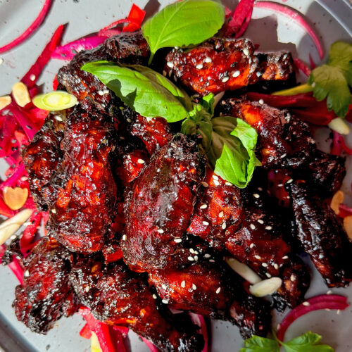 Dark red glazed crispy sticky honey soy chicken wings piled on a plate with sesame seeds, fresh herbs, beetroot salad, red onion, and toasted almonds. These air fryer chicken wings are healthy, dairy-free, high-protein, naturally sweetened, smoky, caramelised and sweet, savoury, tender inside, crispy around the edges, and coated in a next-level sticky secret glaze.