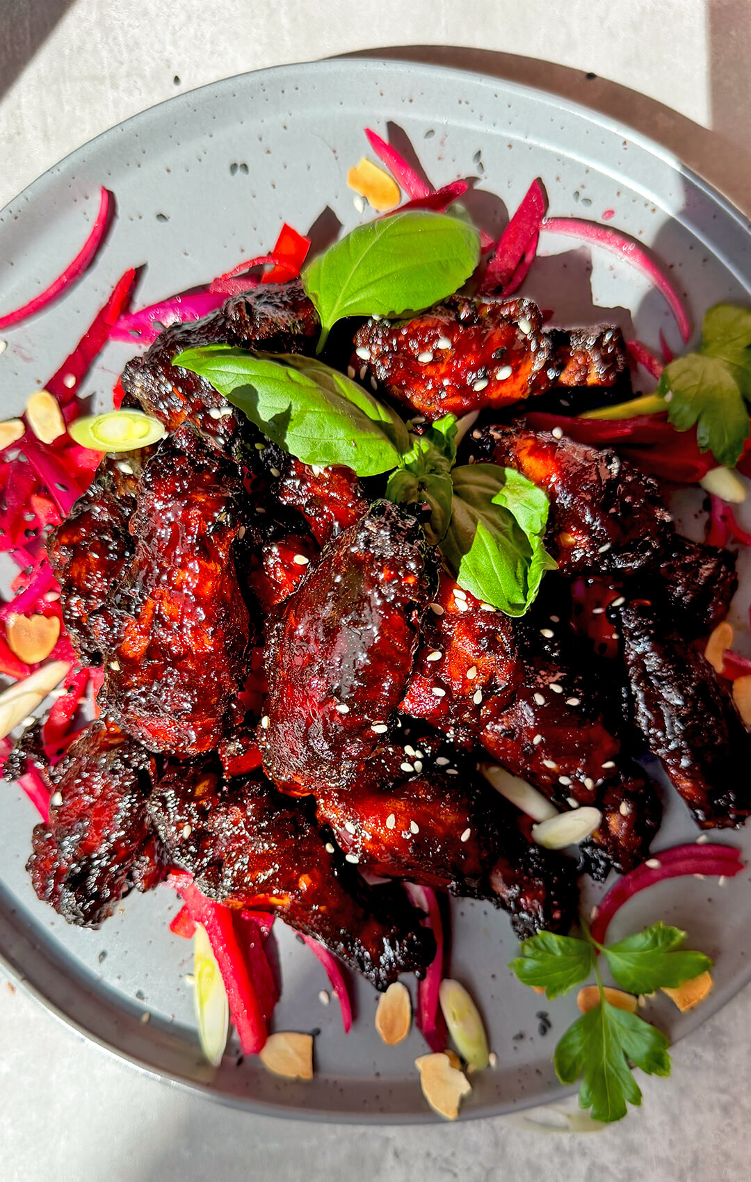 Dark red glazed crispy sticky honey soy chicken wings piled on a plate with sesame seeds, fresh herbs, beetroot salad, red onion, and toasted almonds. These air fryer chicken wings are healthy, dairy-free, high-protein, naturally sweetened, smoky, caramelised and sweet, savoury, tender inside, crispy around the edges, and coated in a next-level sticky secret glaze.