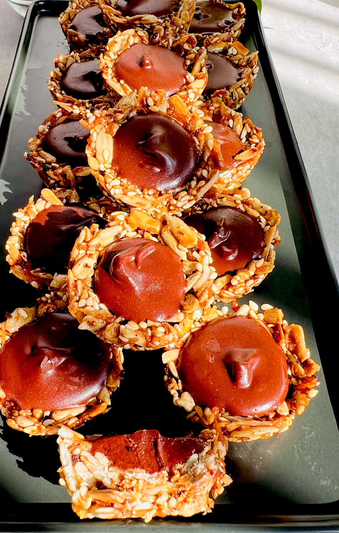No-Oven Chocolate Tahini Seed Cups on a black serving plate, showing crunchy golden toasted seed cups filled with silky, glossy dark chocolate tahini, plus one cut cup showing the creamy centre. These healthy homemade chocolate cups are paleo, gluten-free, nut-free, dairy-free, oil-free, refined sugar-free, delicious, satisfying, and perfect for a wholesome dessert, snack, or lunch box treat.