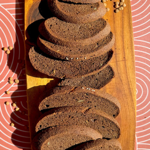 Sliced rustic brown lentil bread on a wooden board with brown lentils, seeds, cocoa powder, and caraway in the background. This gluten-free, flourless bread is yeast-free, egg-free, high in fibre, protein-rich, easy to make at home, and has a delicious dark rye-style flavour and look without tasting like lentils.
