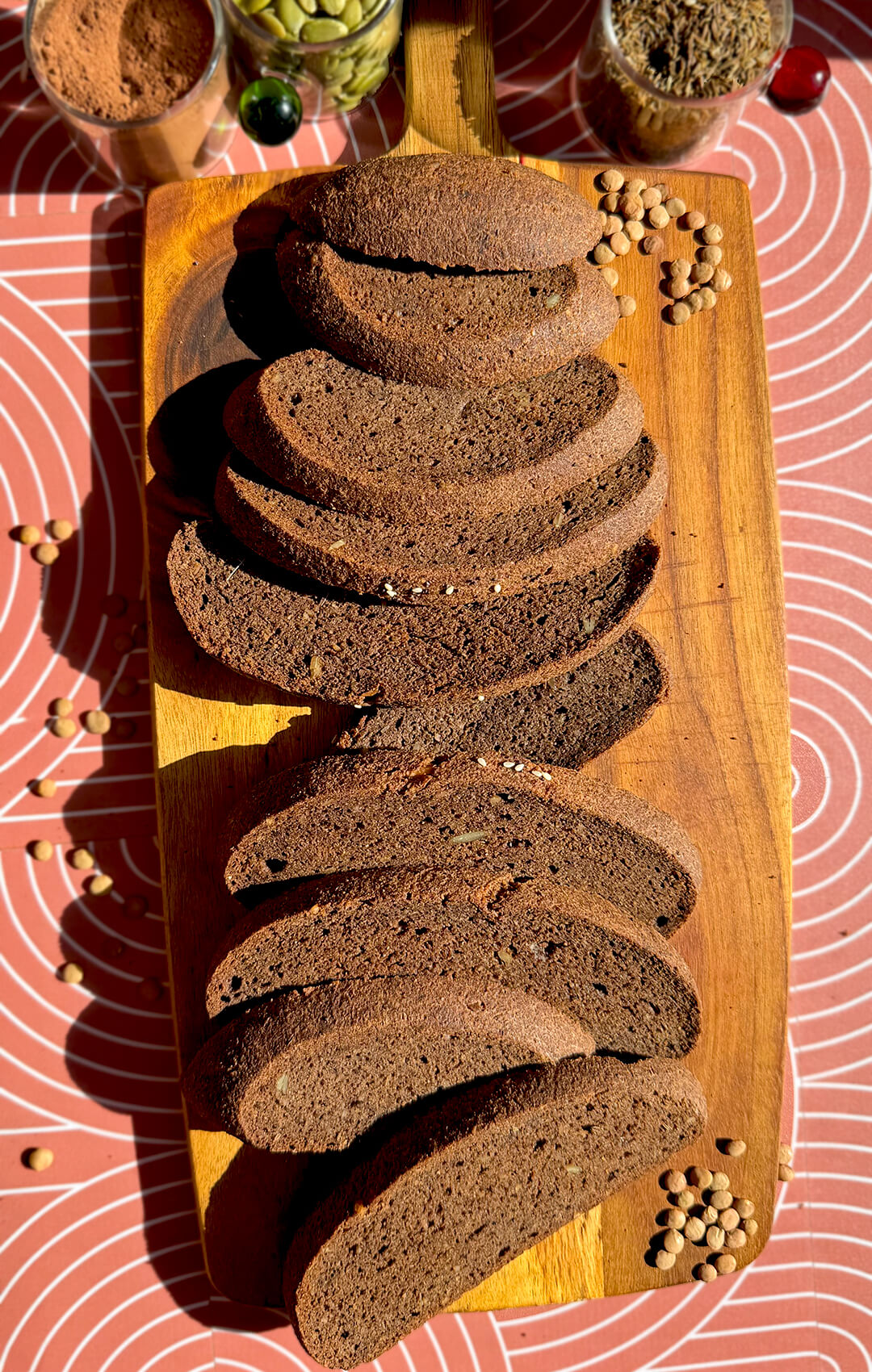 Sliced rustic brown lentil bread on a wooden board with brown lentils, seeds, cocoa powder, and caraway in the background. This gluten-free, flourless bread is yeast-free, egg-free, high in fibre, protein-rich, easy to make at home, and has a delicious dark rye-style flavour and look without tasting like lentils.