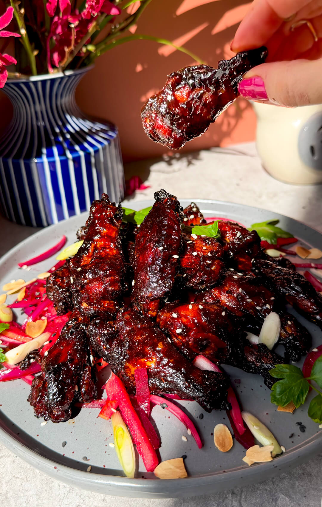 Crispy sticky honey soy air fryer chicken wing held above a serving plate, coated in a glossy dark red secret glaze with crispy caramelised edges. These healthy air fryer chicken wings are naturally sweetened with honey, dairy-free, high-protein, smoky, sweet, savoury, slightly spicy, tender and juicy inside, and deliciously sticky for an easy homemade dinner, party food, or movie night recipe.