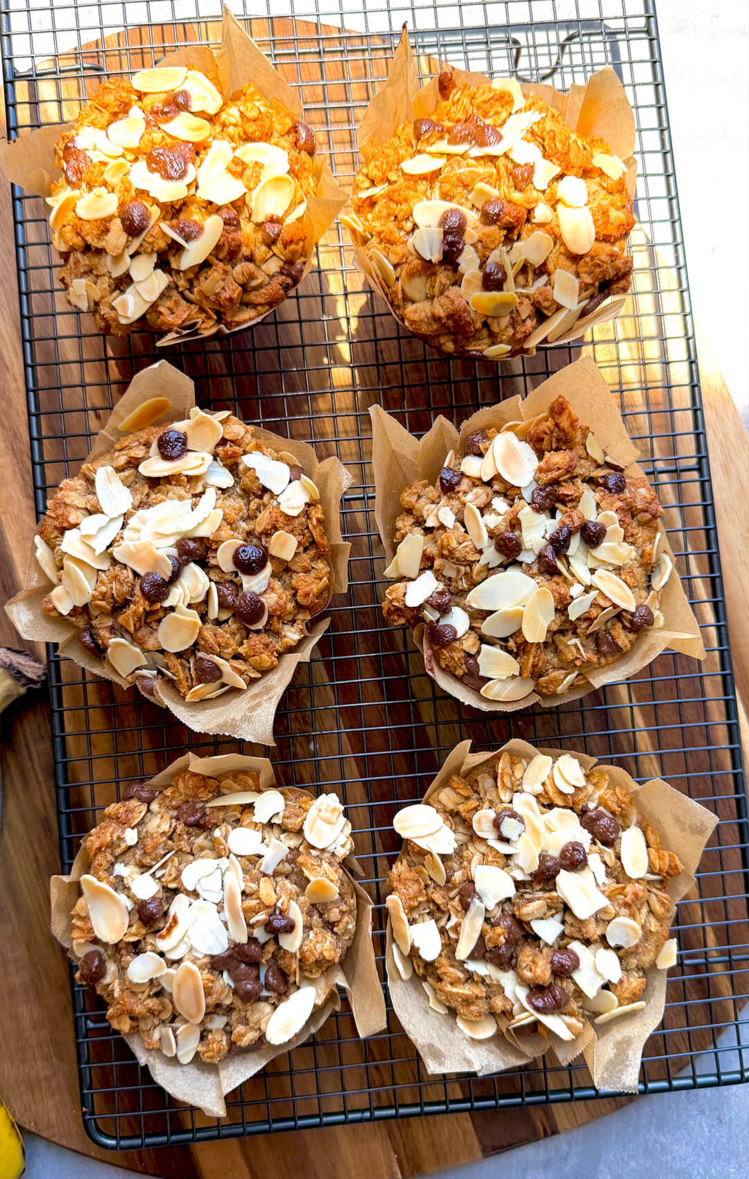 Gluten-free jumbo banana bread crumble muffins cooling on a wire rack, topped with golden chunky oat almond crumble, sliced almonds, and sugar-free chocolate chips. These soft, delicious, bakery-style banana muffins are dairy-free, refined sugar-free, naturally sweetened, satisfying, and perfect for a healthy breakfast, snack, or easy meal prep at home.