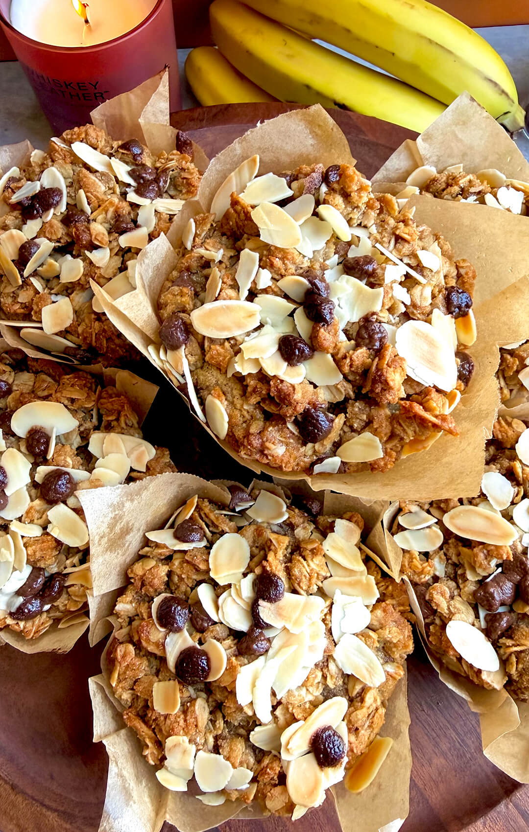 Bakery-style gluten-free banana bread crumble muffins topped with chunky oat almond crumble, sliced almonds, and sugar-free chocolate chips, served with ripe bananas in the background. These soft, golden, delicious muffins are dairy-free, refined sugar-free, naturally sweetened, satisfying, and lovely for breakfast, snacks, lunch boxes, or meal prep.