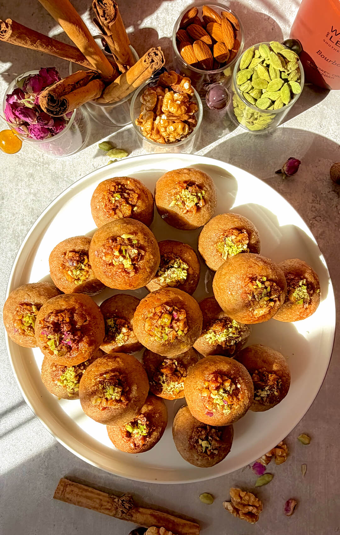 Healthy Baklava Bites shown from above with a golden colour, piled on a white plate with pistachio walnut topping, rose petals, cardamom, cinnamon, walnuts and almonds around the plate. These easy gluten-free, grain-free, dairy-free and refined sugar-free baklava bites are naturally sweetened with honey, golden, nutty, fragrant, crisp outside, moist inside, and perfect for a quick healthy homemade dessert, sweet snack or afternoon tea treat.