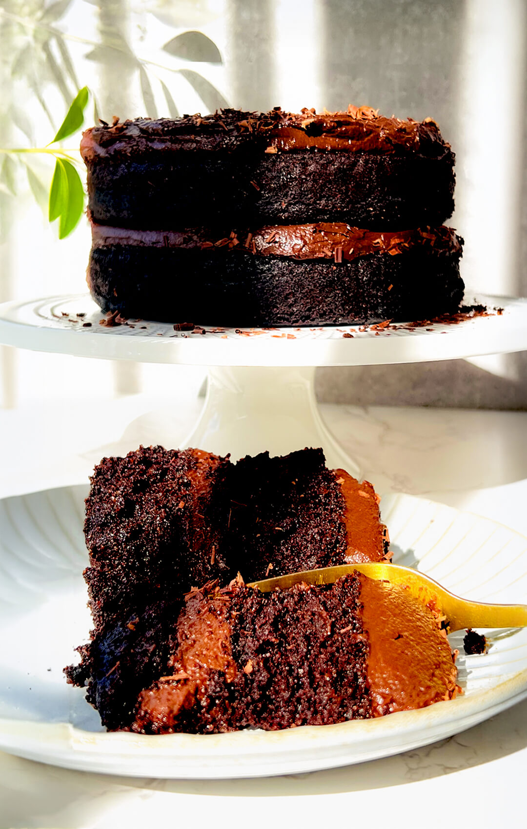 A slice of healthy moist two-layer chocolate cake with a gold fork next to a whole double chocolate paleo gluten-free cake made with almond flour and covered in silky dairy-free ganache, so easy to make at home. It's refined sugar-free, keto option by replacing coconut sugar with brown monkfruite, gluten-free, perfect guilt-free dessert for chocolate lovers.